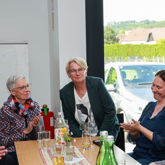 Drei Frauen sitzen um einen Tisch und lächeln. Eine hält eine Blume. Ein Auto ist draußen vor dem Fenster geparkt.