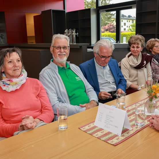 Eine Gruppe älterer Menschen sitzt an einem Tisch mit Namenskarten und Blumen, einer hält ein Handy. Der Raum hat eine rote Wand, ein Regal und ein Fenster mit Bäumen.