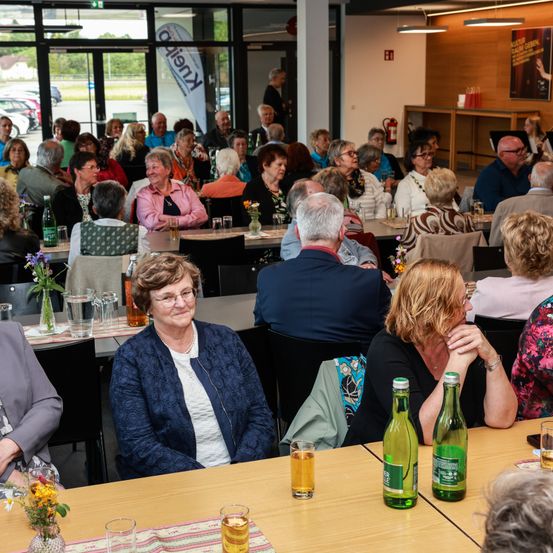 Ein Speisesaal mit mehreren älteren Menschen, die an Tischen sitzen, einige mit Brille und Halsketten. Flaschen mit Wein und Bier stehen auf den Tischen. Ein Banner mit 'Klein' auf der linken Seite und ein Feuerlöscher an der Wand.