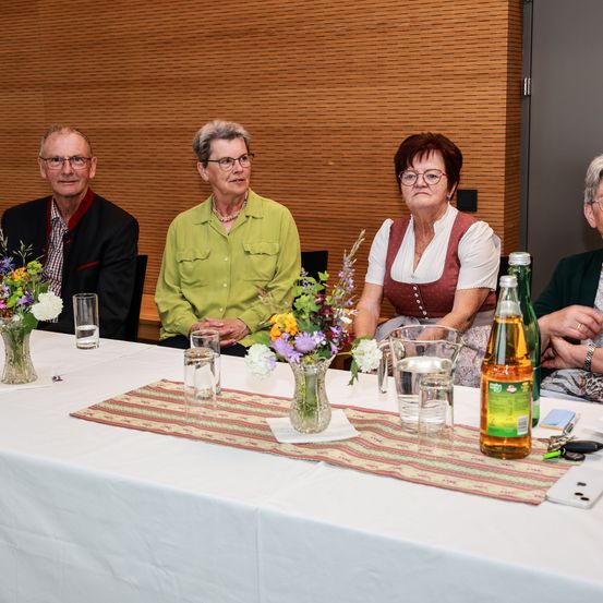 Vier ältere Menschen sitzen an einem Tisch mit Blumen in Vasen, Gläsern mit Wasser und einer Karaffe. Sie sehen ernst aus, wahrscheinlich bei einer Besprechung. Dahinter befindet sich eine Holzwand und eine offene Tür.