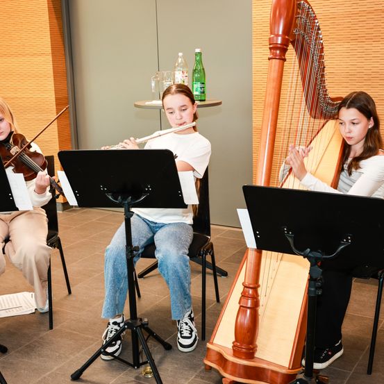 Drei junge Mädchen sitzen auf Stühlen und spielen jeweils ein Musikinstrument. Das Mädchen links spielt Geige, die in der Mitte Flöte und das rechts Harfe. Notenständer stehen vor ihnen.