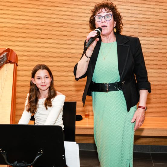 Eine Frau steht neben einem Mädchen, das eine Harfe spielt, und hält ein Mikrofon, möglicherweise beim Singen. Dahinter ist eine Holzwand mit einer Bank und einem Glas mit Flüssigkeit zu sehen.