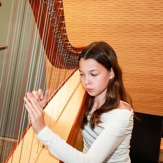 Ein junges Mädchen spielt eine Harfe in einem Raum mit einer Holzwand. Sie konzentriert sich auf ihre Aufführung, ihre Hände bewegen sich über die Saiten.