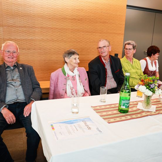 Eine Gruppe älterer Menschen sitzt an einem Tisch, lächelt und scheint eine Mahlzeit zu genießen. Der Tisch hat Blumen, Gläser und eine Flasche. Hinter ihnen ist eine Holzwand und eine graue Tür zu sehen.