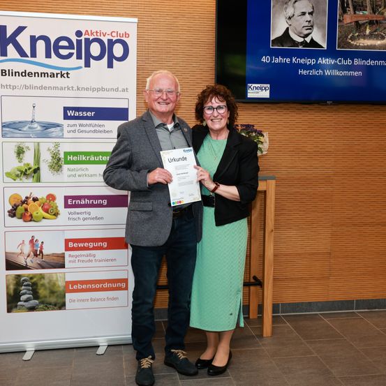 Ein älterer Mann und eine Frau stehen nebeneinander und halten ein Zertifikat. Dahinter befindet sich ein Banner, das die Gesundheitsvorteile von Kneipp bewirbt.