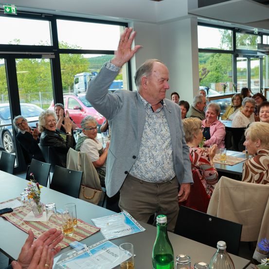 Ein älterer Mann winkt einem sitzenden Publikum zu. Sie befinden sich in einem Essbereich mit großen Fenstern, Autos sind draußen sichtbar. Der Mann lächelt, das Publikum klatscht.
