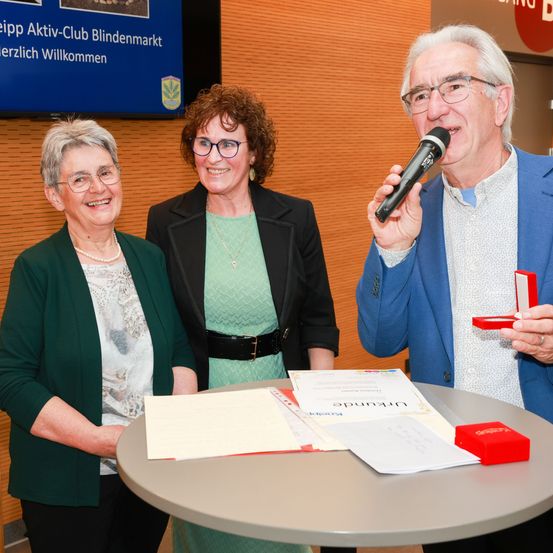 Drei Erwachsene stehen um einen Tisch mit Zertifikaten. Ein Mann hält ein Mikrofon und eine Frau eine Schachtel. Der Mann lächelt und spricht in das Mikrofon. Hinter ihnen steht ein Schild mit der Aufschrift 'Leipzig Aktiv-Club Blindenmarkt herzlich Willkommen.'
