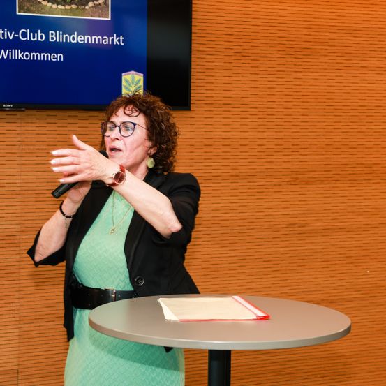 Eine Frau spricht in ein Mikrofon an einem Rednerpult vor einem Schild, das 'Willkommen beim Blindenmarkt Aktiv-Club' besagt.