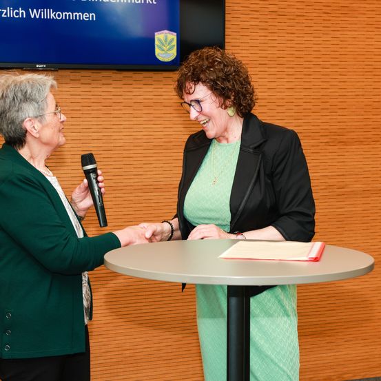 Zwei Frauen interagieren an einem Tisch, eine hält ein Mikrofon. Die Frau in Grün lächelt und schüttelt die Hand der Frau in Schwarz. Dahinter zeigt ein Bildschirm eine Willkommensnachricht an.