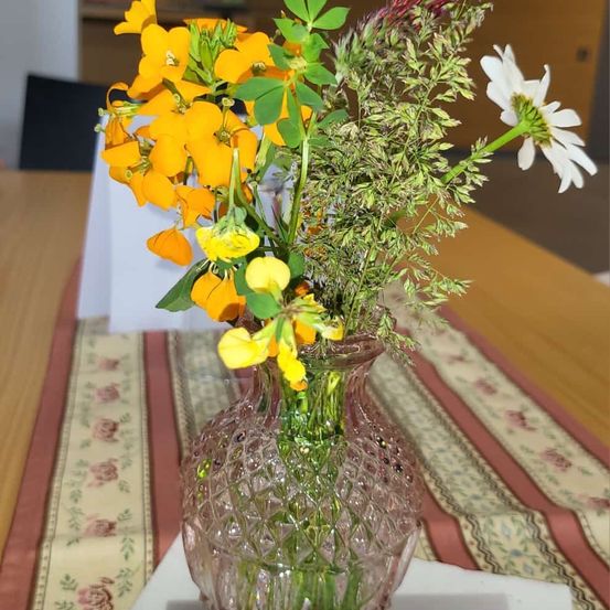 Eine klare Glasvase mit gelben und weißen Blumen steht auf einem Tisch, der mit einem Blumentischtuch bedeckt ist.