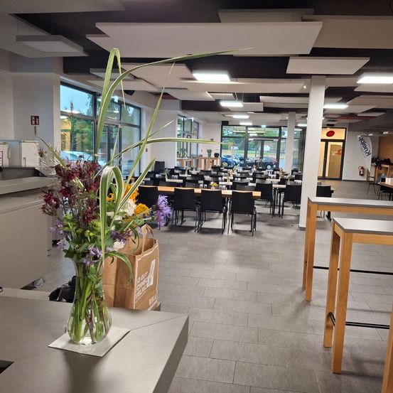Ein leerer Speisesaal mit Tischen, Stühlen und einer Blumenvase. Der Saal hat große Glasfenster, eine weiße Decke und geflieste Böden.