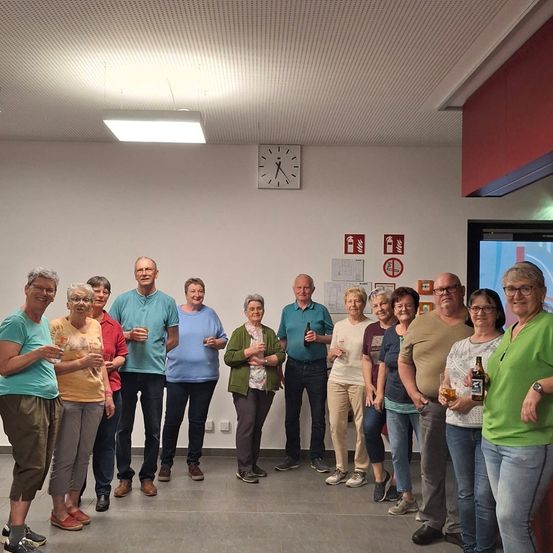 Eine Gruppe von Menschen in Freizeitkleidung steht in einem Raum zusammen und hält Getränke in den Händen. Sie scheinen ein Ereignis zu feiern. Eine Uhr und Plakate sind an der Wand angebracht.