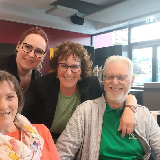 Vier Menschen lächeln auf einem Foto, wahrscheinlich in einem Restaurant. Die Frau auf der linken Seite trägt ein Halstuch, während der Mann auf der rechten Seite ein grünes Hemd trägt. Die beiden Frauen in der Mitte tragen Brillen und haben ihre Arme umeinander geschlungen.