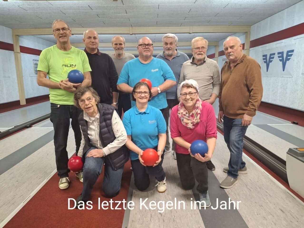 Eine Gruppe von Menschen posiert für ein Foto in einer Bowlingbahn. Sie halten Bowlingkugeln in den Händen und lächeln. Die Worte 'Das letzte Kegeln im Jahr' sind auf dem Boden geschrieben.