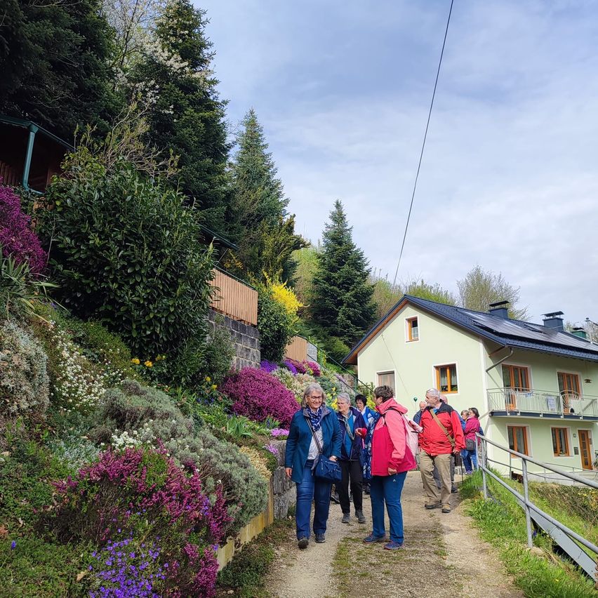 Eine Gruppe von Menschen geht auf einem von bunten Pflanzen umgebenen Pfad. Sie gehen auf ein Haus mit Solarpaneelen auf dem Dach zu.