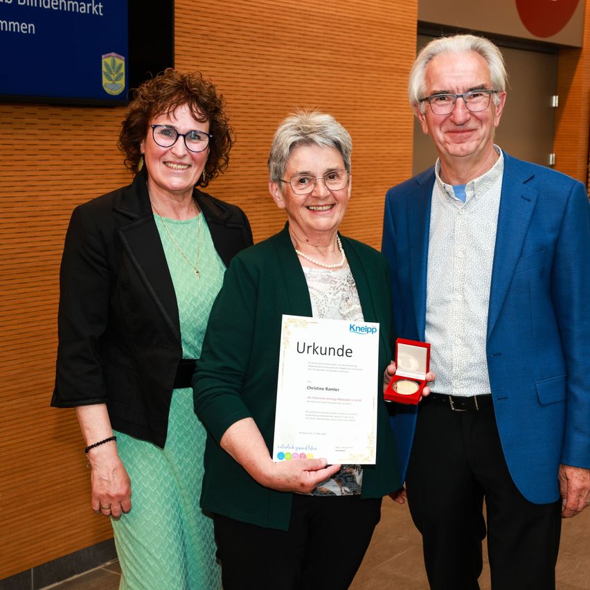 Three people are standing close together. The woman in the middle holds a certificate. They are all smiling. Behind them is a wooden wall with a blue sign.