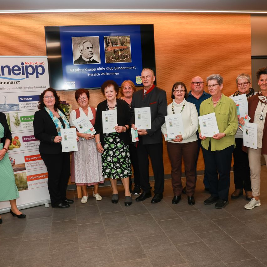 A group of adults poses for a photo in front of a banner. They are holding certificates. In the background, there is a screen with a picture of a man.