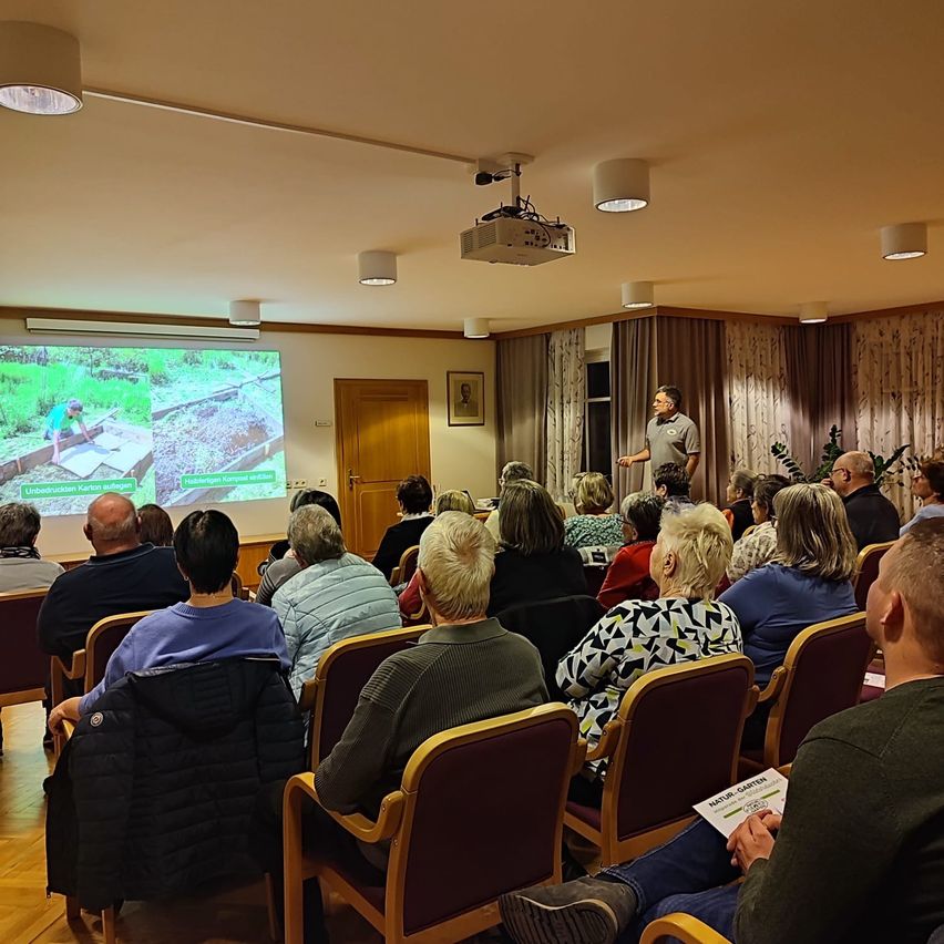 Ein Mann steht vor einer Projektionsleinwand und hält eine Präsentation für ein Publikum in einem Konferenzraum. Das Publikum sitzt in Reihen von Stühlen.