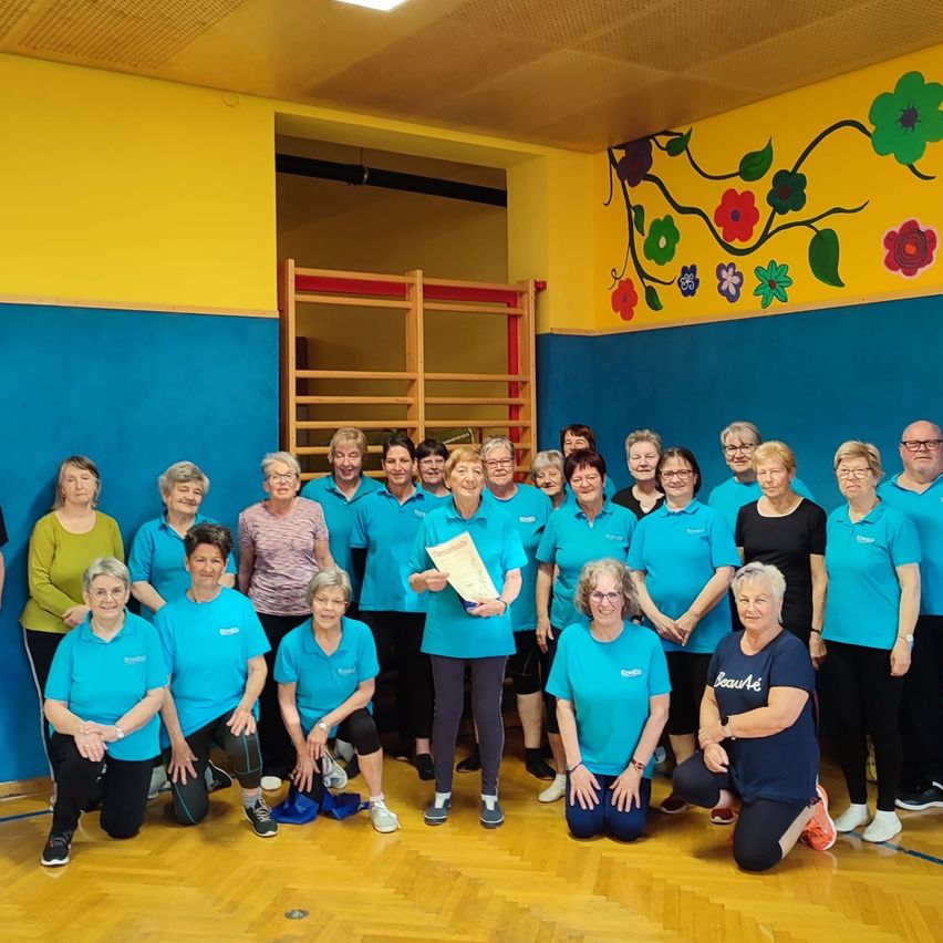 Eine Gruppe von Frauen in blauen Shirts und schwarzen Hosen posiert für ein Foto in einem Fitnessstudio mit einer gelben und blauen Wand und einer Wandmalerei. Eine Frau hält ein Papier, und eine Frau in einem schwarzen Shirt sagt 'Beaute'.