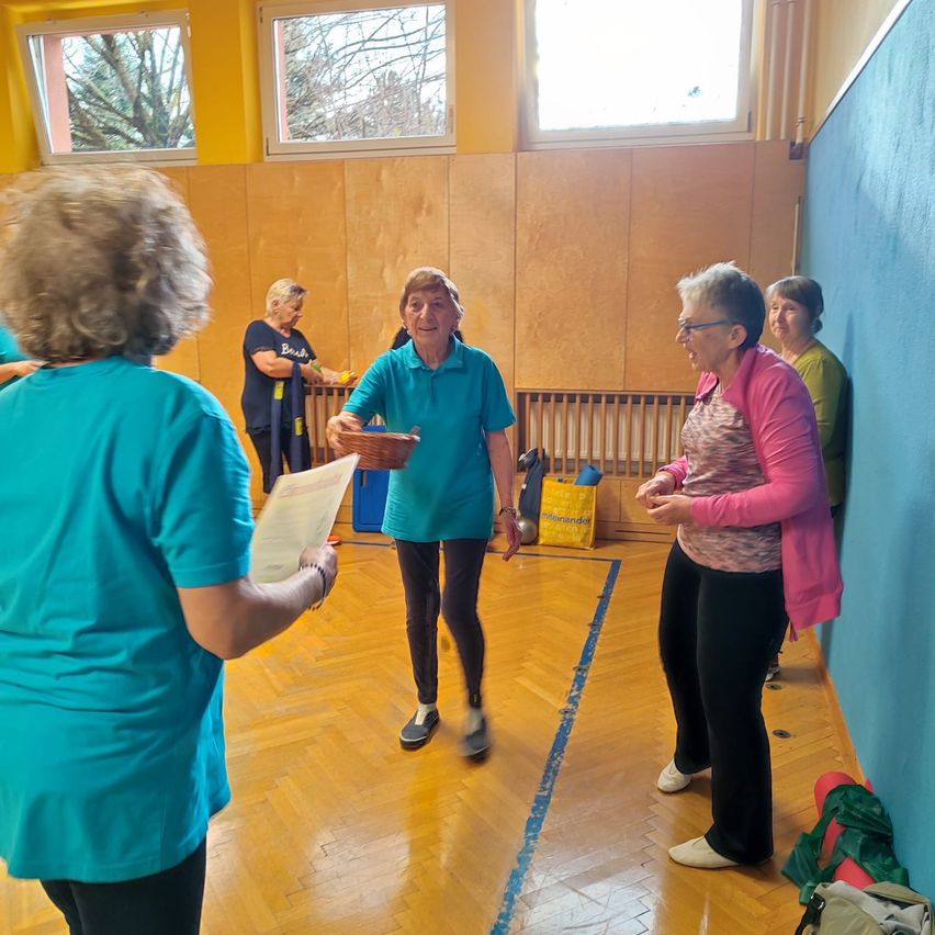 Eine ältere Frau mit einem Korb steht zwischen zwei Frauen in einer Turnhalle, während andere im Hintergrund sind. Der Raum hat Holzböden, gelbe Wände und Fenster.