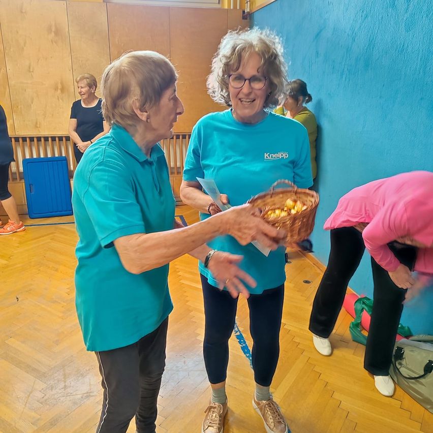 Zwei ältere Frauen stehen in einem Innenraum, eine hält einen Korb. Eine Frau trägt eine Brille und ein Armband. Eine andere Frau beugt sich herunter.