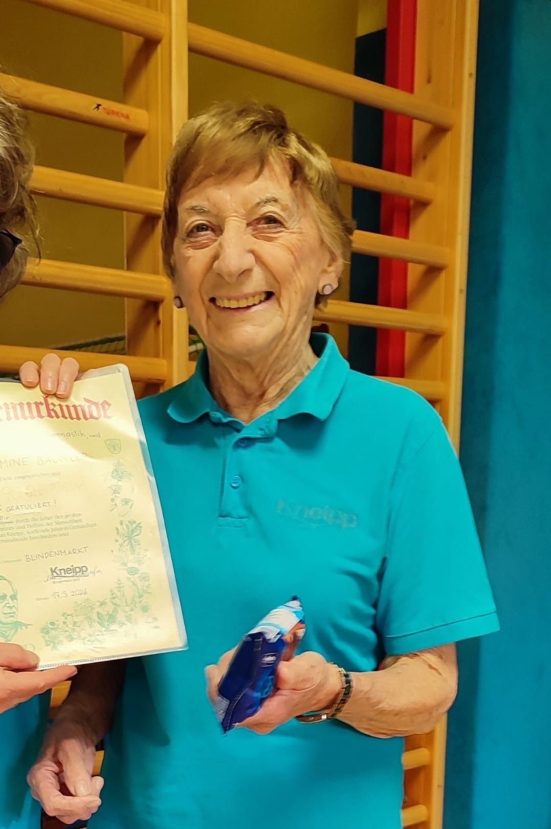 Eine ältere Frau in einem blauen Shirt hält einen Zertifikat und lächelt. Das Zertifikat ist weiß mit Text und einem Siegel. Die Frau steht neben einer anderen Person, die ein blaues Objekt hält. Hinter ihnen steht eine Holzstruktur mit einem gelben und roten Rand.