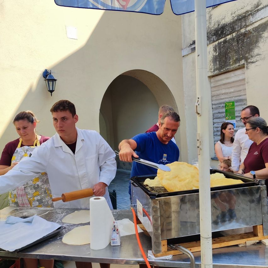 Eine Gruppe von Menschen kocht im Freien, wobei ein Mann in einem blauen Shirt Teig auf einem Grill wendelt. Ein anderer Mann in einem weißen Mantel rollt Teig auf einem Tisch.