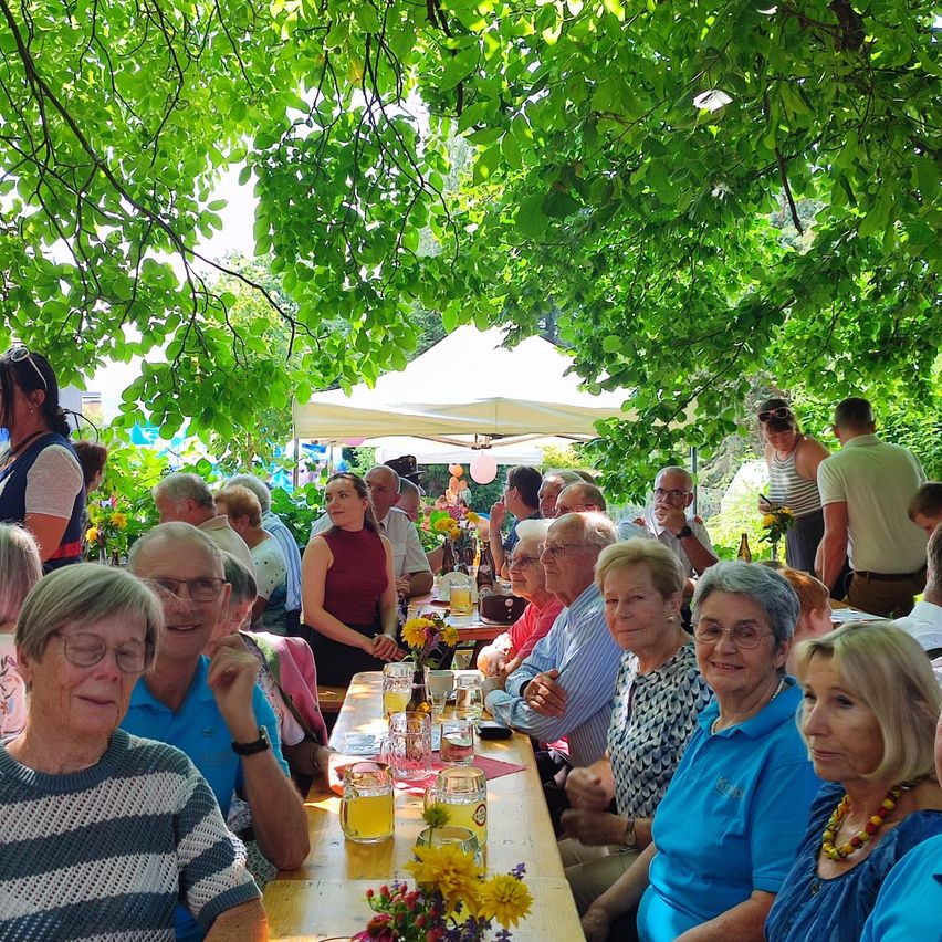 Eine Gruppe älterer Menschen sitzt um einen langen Tisch unter einem Baum herum, wahrscheinlich bei einer Gartenparty. Einige tragen Brillen und lächeln, während andere in Gespräche vertieft sind. Auf dem Tisch stehen Blumen, Gläser und Flaschen. Im Hintergrund befinden sich Zelte und Bäume.