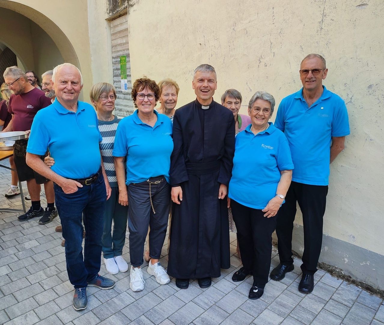 Eine Gruppe von Menschen, einschließlich eines Priesters, steht lächelnd vor einem Gebäude für ein Foto.