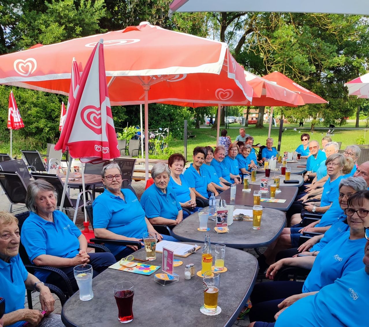 A group of older adults in blue shirts sit under umbrellas at an outdoor cafe, with drinks and snacks on the tables.