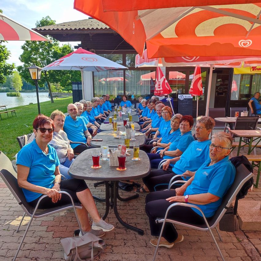 A group of elderly people are sitting around a table under an umbrella on a patio. They are all wearing blue shirts. There are several glasses of beer on the table. Behind them is a building with a glass door and windows.