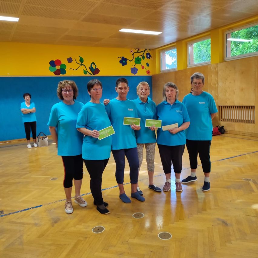 Five women in blue shirts pose for a photo in a gym with a yellow and blue wall, holding certificates. Another woman stands near a bag.