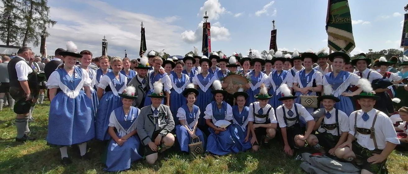 Eine Gruppe von Menschen in traditionellen Kostümen, einige mit Fächern und Körben, posiert für ein Foto auf einer Rasenfläche mit Fahnen und Bannern im Hintergrund unter einem bewölkten Himmel.