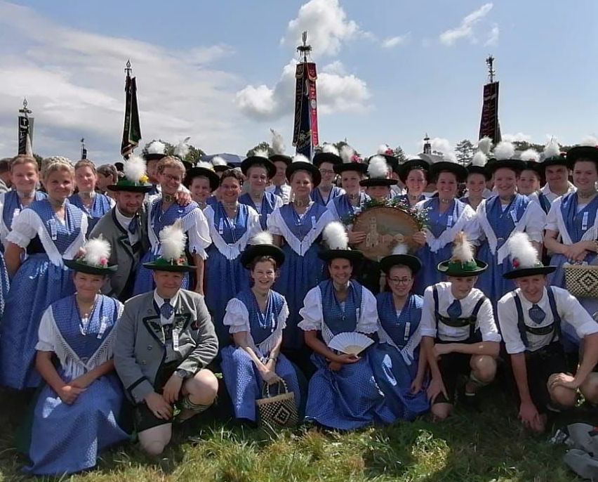 Eine Gruppe von Menschen in traditionellen Kostümen, einige mit Fächern und Körben, posiert für ein Foto auf einer Rasenfläche mit Fahnen und Bannern im Hintergrund unter einem bewölkten Himmel.