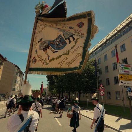 Eine Gruppe von Menschen in traditioneller bayerischer Kleidung marschiert in einem Umzug. Ein Banner mit der Illustration eines Mannes, der ein Fass hält, wird über ihnen gehalten. Gebäude und Flaggen säumen die Straße.