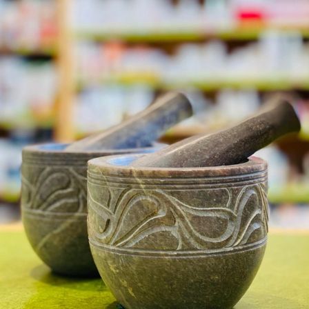 Zwei steinerne Mörser mit Stößeln auf einem grünen Tisch, möglicherweise in einem Geschäft. Die Mörser haben komplizierte Designs. Im Hintergrund befindet sich ein verschwommener Ladenregal mit Gegenständen.