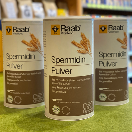 Drei Dosen Raab Spermidin Pulver sind auf einer grünen Oberfläche ausgestellt. Die Dosen sind mit dem Markennamen Raab beschriftet und enthalten Weizenkörner.