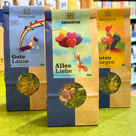 Drei Tüten Bio-Kräutertee sind ausgestellt. Die erste Tüte ist blau mit einem Regenbogen und einem Mädchen auf einer Schaukel. Die zweite Tüte ist gelb mit einem Hahn und einem Fuchs, der Luftballons hält. Die dritte Tüte ist braun mit einem Hahn und einer grünen Landschaft.