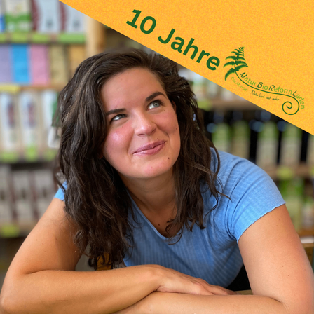 Eine lächelnde Frau mit lockigem Haar in einem blauen Shirt posiert in einem Geschäft mit Regalen von Produkten im Hintergrund. Das Schild oben sagt '10 Jahre'.