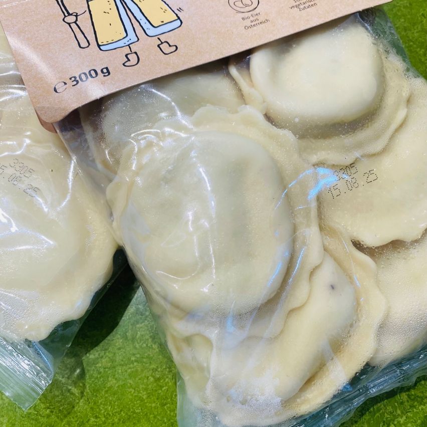 Packung mit gefrorenem Ravioli in Plastiktüten, mit einem Etikett, das das Gewicht von 300 g angibt, auf einer grünen Oberfläche aufbewahrt.