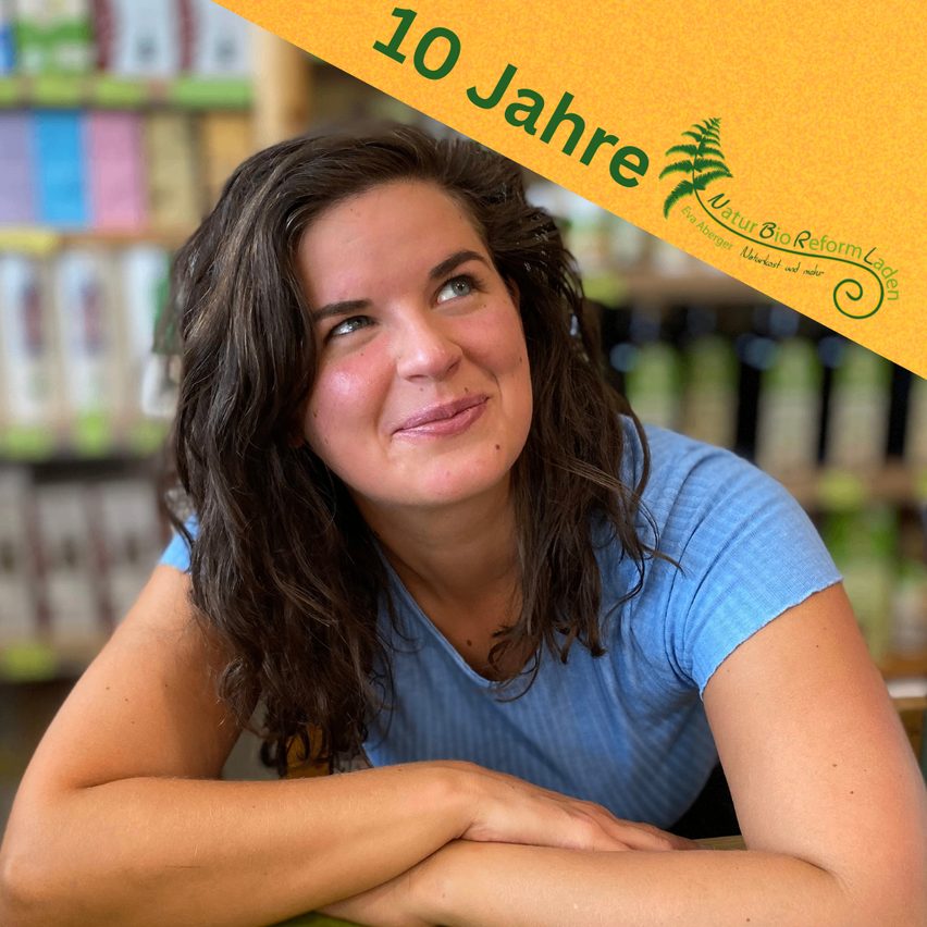 Eine Frau mit lockigem Haar und einem blauen Shirt lächelt für die Kamera, mit Regalen voller Produkte im Hintergrund. Ein Overlay zeigt '10 Jahre' in Deutsch.