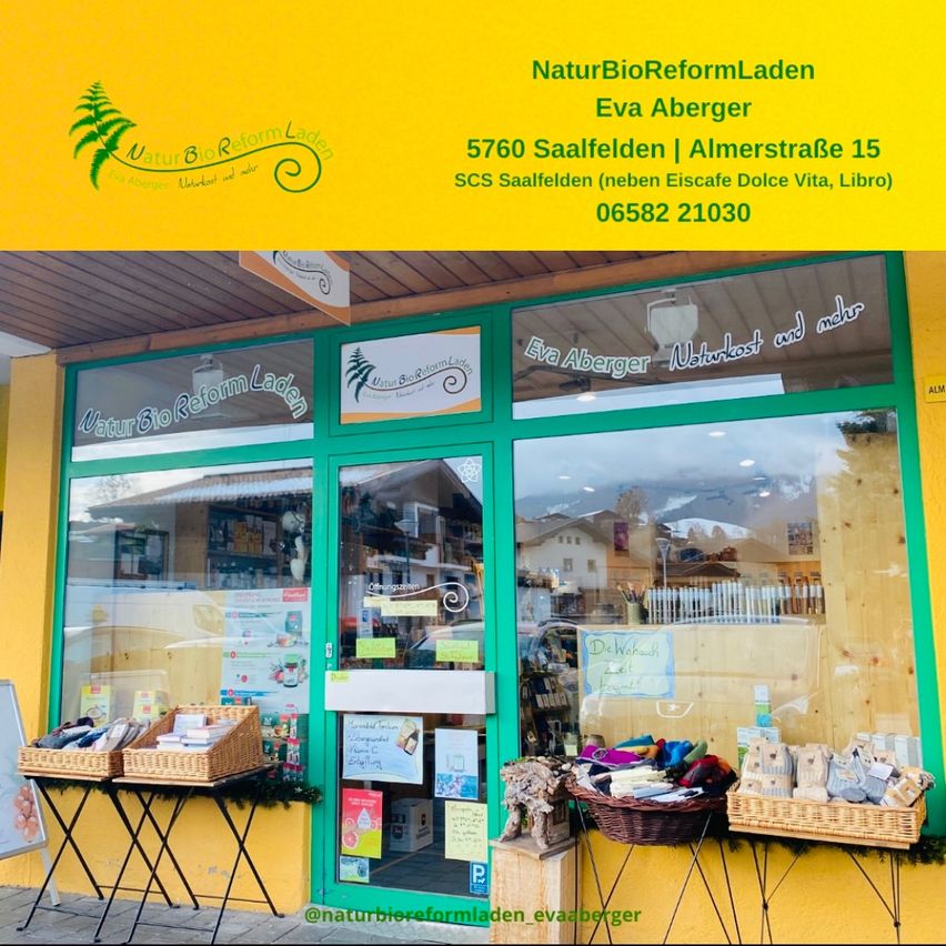 Ein Ladengeschäft mit gelben Wänden und grünem Rahmen zeigt eine Glastür und ein Fenster. Zwei Körbe mit Produkten sind draußen ausgestellt, mit einem Baumlogo oben. Das Schild lautet NaturBioReformLaden und Eva Aberger.