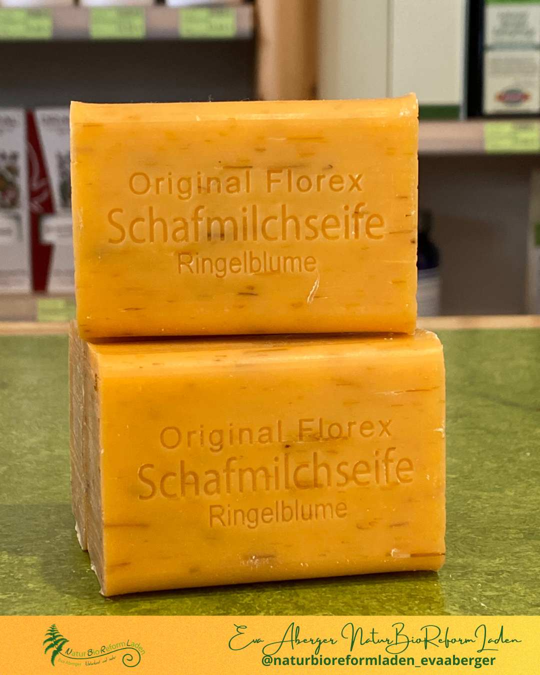 Zwei gelbe Seifenstücke sind aufeinandergestapelt. Die Seife ist mit 'Original Florex Schafmilchseife Ringelblume' beschriftet. Die Seife liegt auf einer grünen Oberfläche. Hinter der Seife befindet sich ein unscharfer Hintergrund mit einem Regal und einigen Kisten.