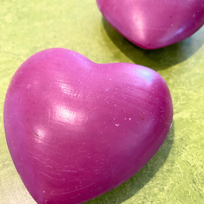 Zwei lila herzförmige Objekte liegen auf einer grünen Oberfläche. Eines ist vorne und etwas größer, mit einer glatten Textur und einem Schatten. Das andere ist dahinter und kleiner.