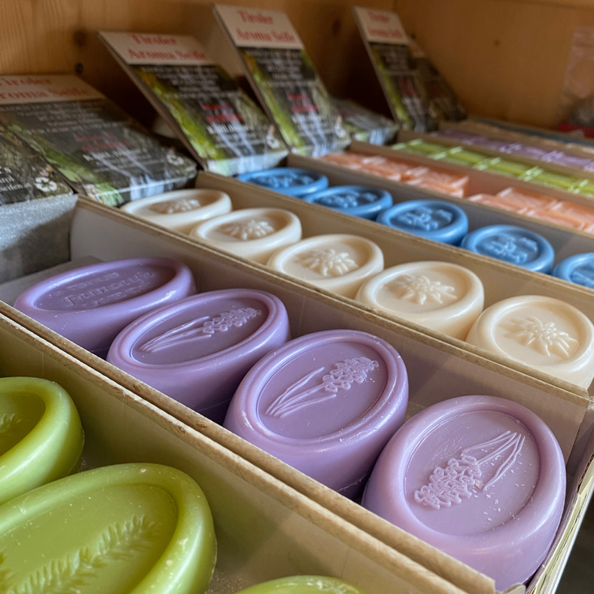 Ein Holzregal hält mehrere Seifenschachteln. Die Seifen sind in verschiedenen Farben und Formen, einige mit Blumendesigns. Die Schachtel ist geöffnet und zeigt mehr Seifen im Inneren.