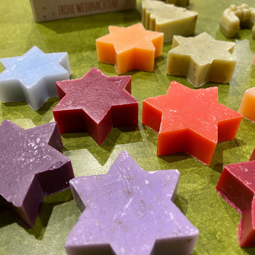 Mehrere sternförmige Seifen in verschiedenen Farben sind auf einer grünen Oberfläche angeordnet, wobei einige Seifen leicht geschmolzene Ränder haben.