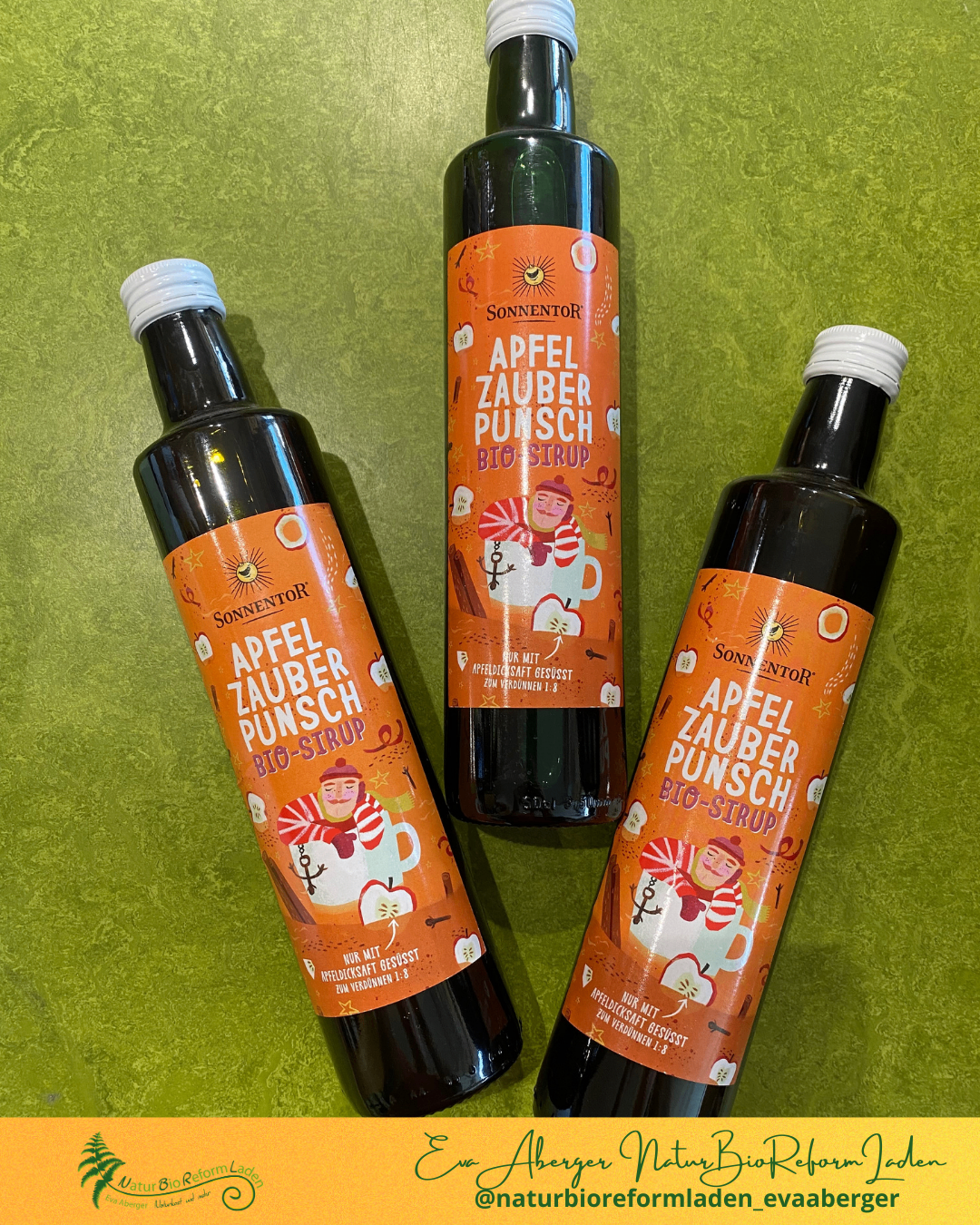 Drei Flaschen biologischer Apfelpunsch-Sirup sind auf einem grünen Hintergrund ausgestellt. Die Flaschen haben orangefarbene Etiketten mit Illustrationen eines Mannes und eines Apfels.