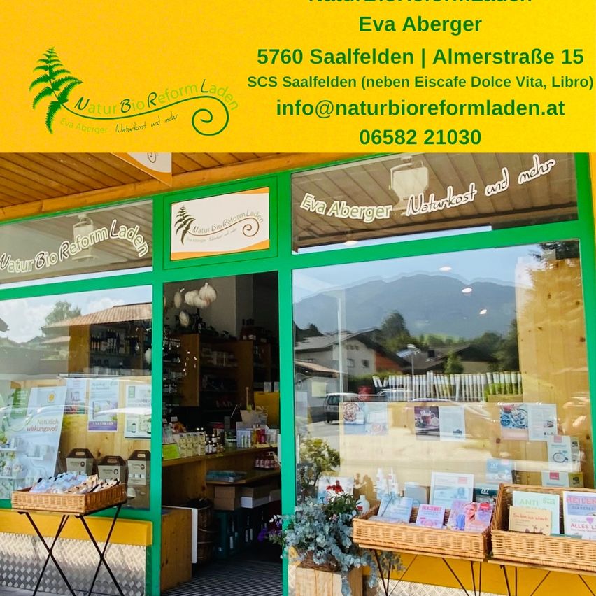 Fassade von Eva Aberger Naturkost, gelegen in Saalfelden. Es hat ein gelbes Schild mit grünen Blättern und Text, eine grüne Tür und Körbe mit Produkten draußen.