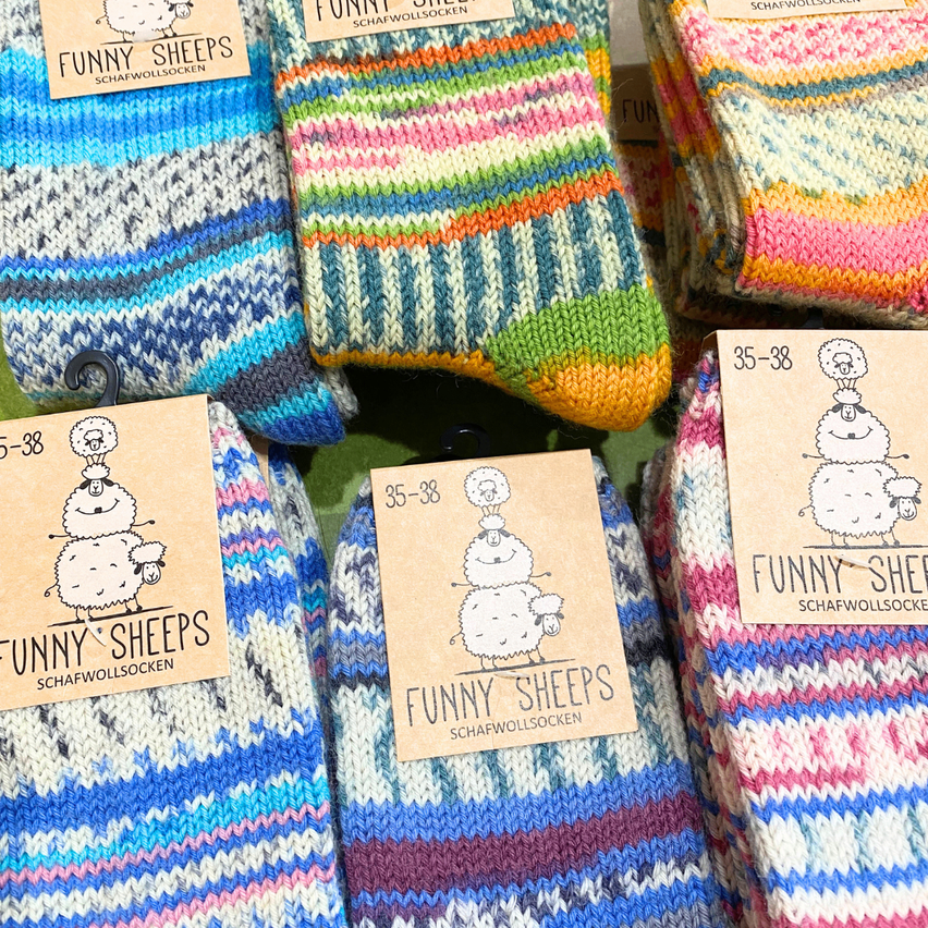 Verschiedene bunte Wollsocken mit der Aufschrift 'Funny Sheeps' werden an einem Kleiderbügel präsentiert. Jede Socke hat einzigartige Streifen und Muster. Die Socken sind in der Größe 35-38 erhältlich.