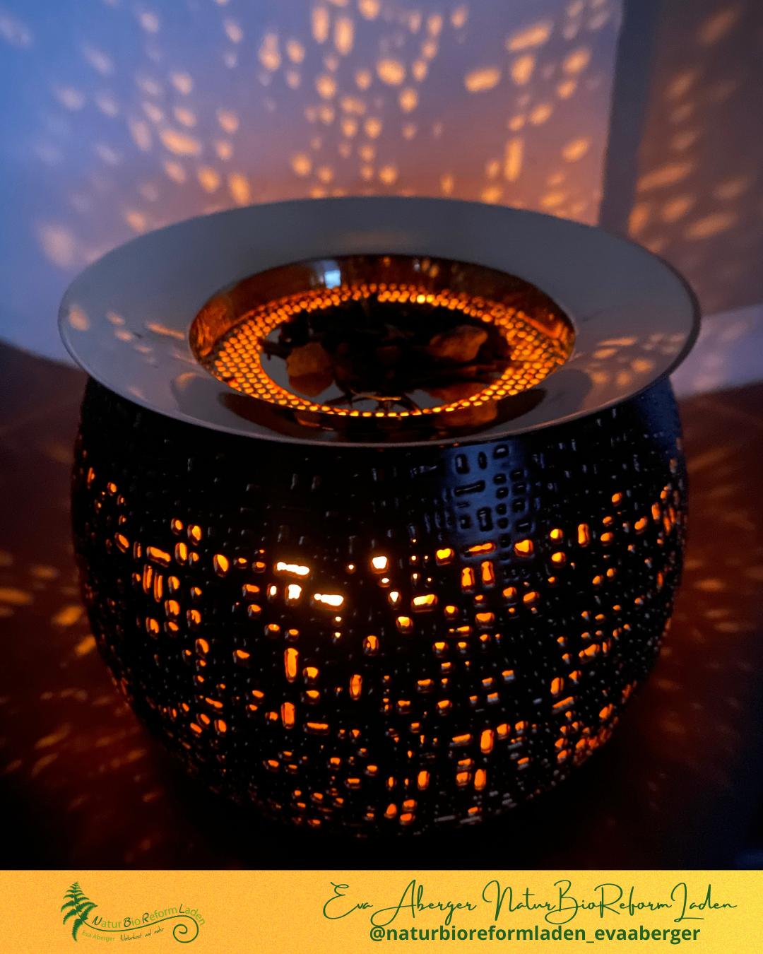 Ein dekorativer schwarzer und goldener Kerzenwärmer ist beleuchtet und wirft ein warmes Licht und Schatten an die Wand.